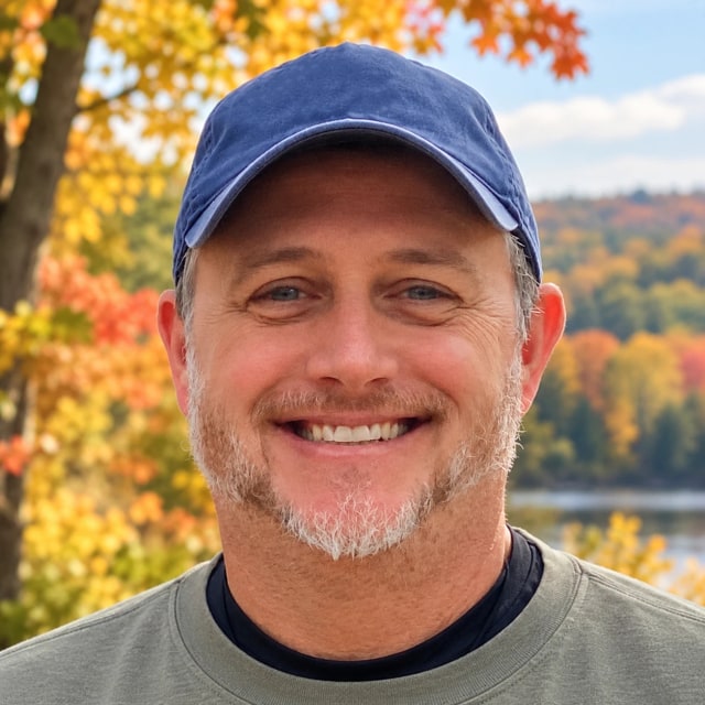 Brian J. Mueller smiling in a navy ball cap, autumn maples and a lake behind him
